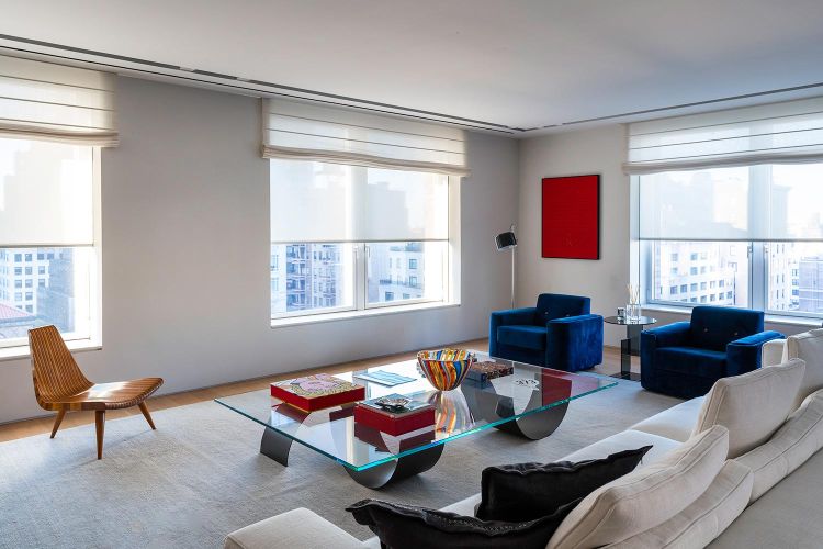 Bright modern living room with a large white sofa, glass coffee table, and two blue armchairs