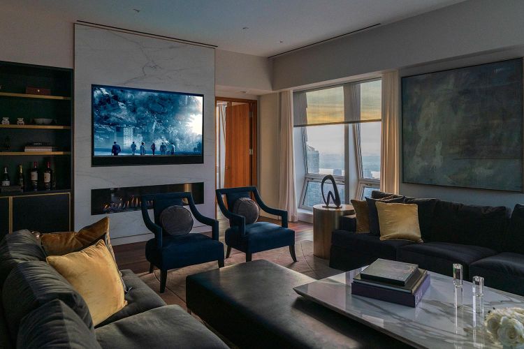 Modern living room with dark sofas, marble fireplace with tv, and large window at dusk