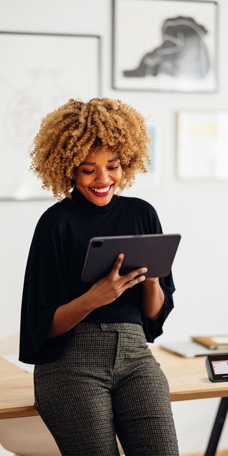 Smiling woman with curly hair using a tablet while leaning against a wooden office desk