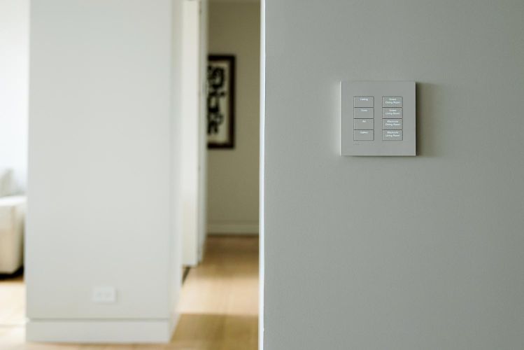 Minimalist smart home keypad mounted on a grey wall with a blurred hallway background