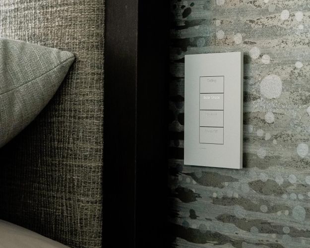 Angled close-up of a wall keypad for lighting and shade automation beside a bed frame