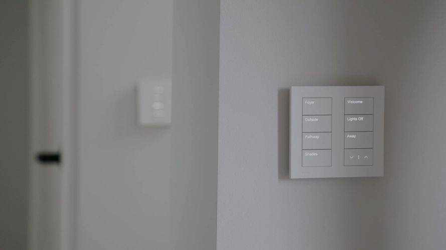 Close-up of a white smart home keypad with labeled backlit buttons mounted on a grey wall
